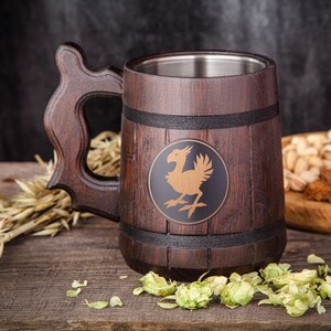 Könnte beinhalten: Ein Bierkrug aus Holz mit dekorativem Griff und Edelstahl-Innenraum. Der Krug hat ein rundes Emblem mit einem goldenen Vogeldesign. Hopfen, Getreide und Snacks sind um den Krug herum auf einer Holzoberfläche angeordnet.
