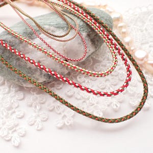 Puede incluir: Primer plano de varios collares de cordón trenzado de varios tamaños. Los cordones presentan patrones rojos, blancos y verdes, algunos en colores lisos. Los collares están dispuestos sobre una piedra gris y un fondo de encaje blanco, lo que sugiere un tema de joyería.