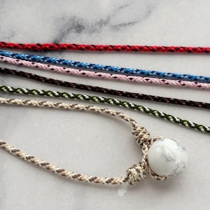 May include: A selection of braided cord necklaces in various colours. One necklace has a white marble bead. The cords are red, blue, pink, brown, green, and a tan and brown pattern. The necklaces are on a light grey surface.