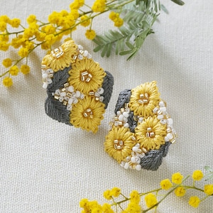 Puede incluir: Un par de pendientes bordados grises y amarillos con un diseño floral. Los pendientes están hechos con pequeñas cuentas y tienen forma de flor.