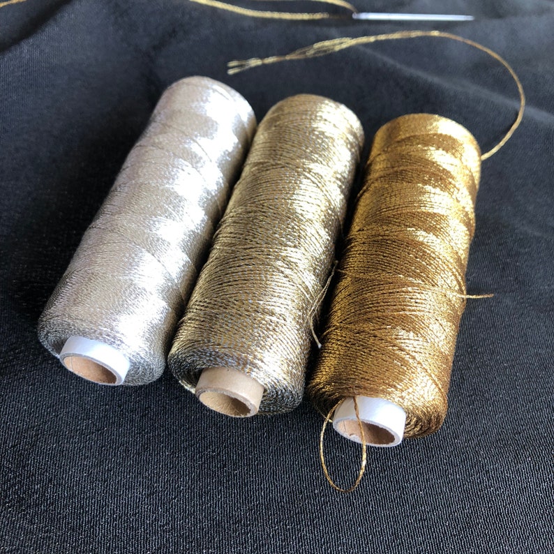 Pu&ograve; includere: Tre bobine di filo metallico, una argentata, una dorata e una bronzea. Le bobine sono appoggiate su un tessuto grigio scuro.