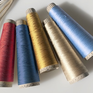 Puede incluir: Cinco carretes de hilo de colores rojo, azul, dorado, beige y azul claro. Los carretes son cilíndricos con base de papel y están dispuestos en fila. Cordones trenzados en beige y negro en la esquina superior izquierda.