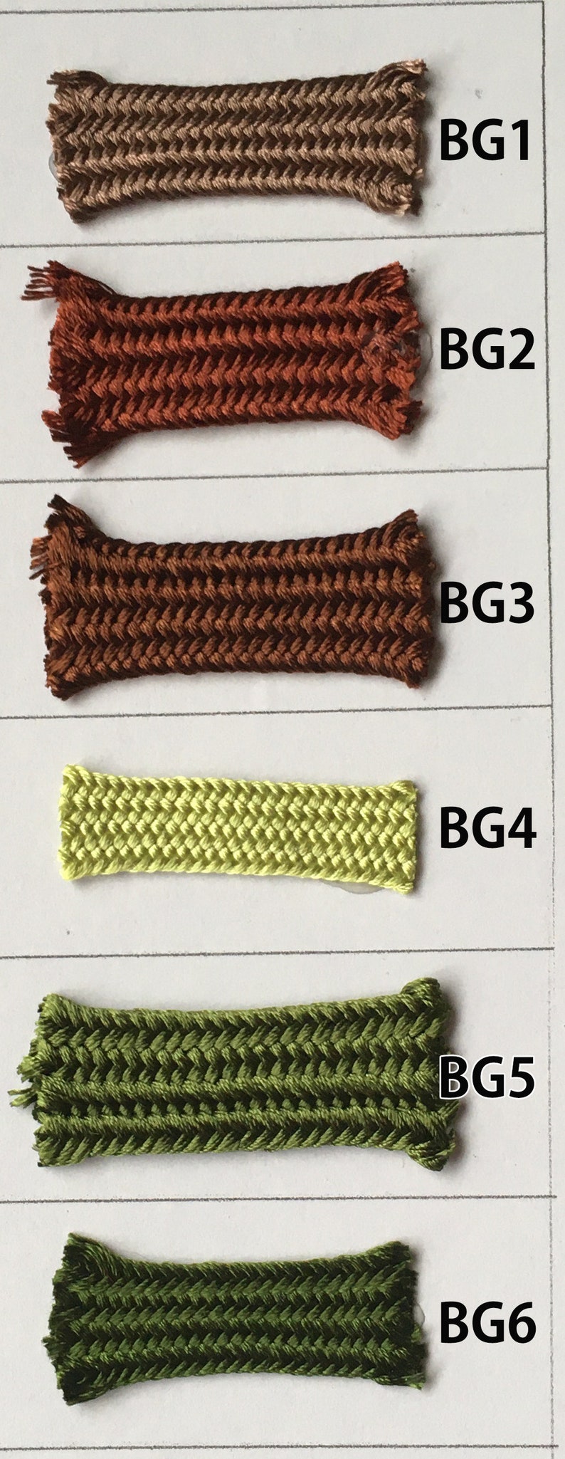 May include: A color chart featuring six samples of braided cord in various shades. The colors range from light brown to dark green. Each sample is labeled with a "BG" followed by a number, indicating different color variations.