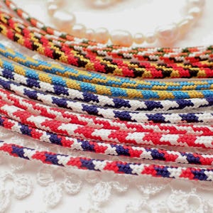 May include: Close-up of several colorful braided cords in red, white, blue, yellow, and black patterns. A strand of white pearls is visible in the background. The cords are likely used for jewelry or crafts.