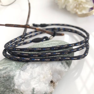 Pode incluir: Correia preta para óculos com detalhes prateados e azuis. A correia é feita de um material tecido e é projetada para segurar os óculos com segurança.