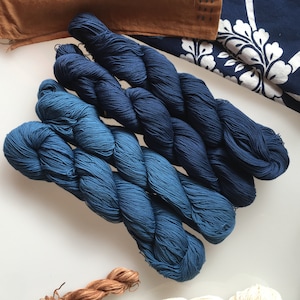 May include: Three skeins of indigo blue yarn, each a slightly different shade, are arranged in a pile. The yarn is soft and textured, and the colours are rich and vibrant.