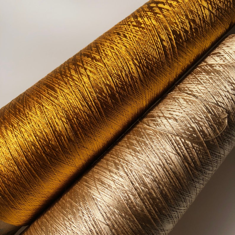May include: Close-up of two spools of metallic thread. One spool is a bright gold color, and the other is a light, silvery gold. The threads are tightly wound, and the texture of the thread is visible.