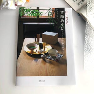 Pode incluir: Um livro branco com o texto japonês "はじめての茶箱あそび" que se traduz como "Primeira vez jogando com a caixa de chá" e "自分だけのひと箱" que se traduz como "Sua própria caixa de chá". A capa do livro apresenta uma foto de uma mesa de madeira com uma bandeja dourada, uma tigela de chá verde, uma tigela pequena branca, uma caixa de madeira e um batedor.