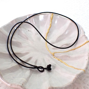 May include: A black cord necklace with a small knot and a decorative element. The necklace is displayed on a white, petal-shaped ceramic dish with gold accents.
