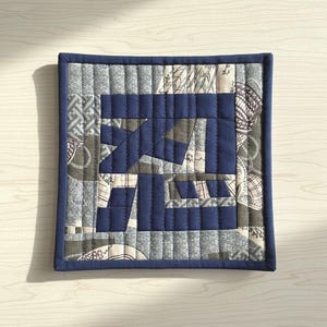 Puede incluir: Textil acolchado cuadrado con un borde azul marino y un diseño central en azul marino y gris. El diseño presenta formas y patrones geométricos, con una mezcla de telas lisas y estampadas. El acolchado crea una superficie texturizada.