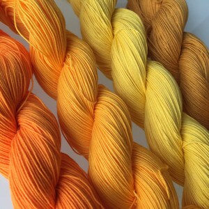 May include: Close-up of several skeins of yarn in shades of orange, yellow, and gold. The yarn is tightly wound and has a smooth texture. The colors transition from bright orange to a deep gold.