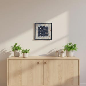 May include: Square wall art featuring a large, dark blue Japanese character on a cream background with a floral pattern. The artwork is framed in a dark blue border and hangs on a white wall. The image also shows a wooden cabinet with plants and decorative jars.