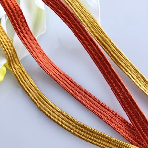 Op de afbeelding: Drie strengen gevlochten trim, één goud en twee oranje, zijn gerangschikt op een wit oppervlak. De trim is geweven met een visgraatpatroon.