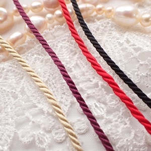 May include: Four strands of twisted cord in beige, burgundy, red, and black are arranged diagonally. The cords feature a smooth, satin-like finish and are positioned over a white lace fabric and a strand of off-white pearls in the background.