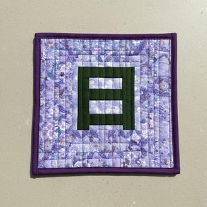 May include: Square quilted coaster with a purple floral pattern and a dark green letter "E" in the center. The coaster has a dark purple border and is likely handmade. The quilting creates a textured surface.