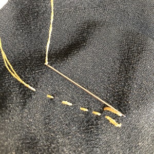 Pu&ograve; includere: Primo piano di un ago e un filo utilizzati per cucire una cucitura su un tessuto nero. Il filo &egrave; di colore giallo chiaro e l'ago &egrave; argentato.