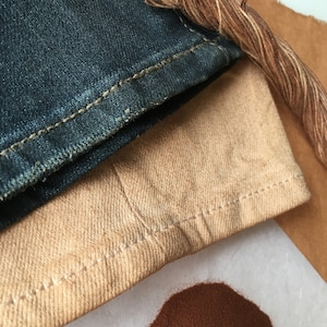 May include: A close-up of a pile of brown dye powder next to a piece of tan fabric and a piece of blue denim fabric. A brown thread is also visible in the image.