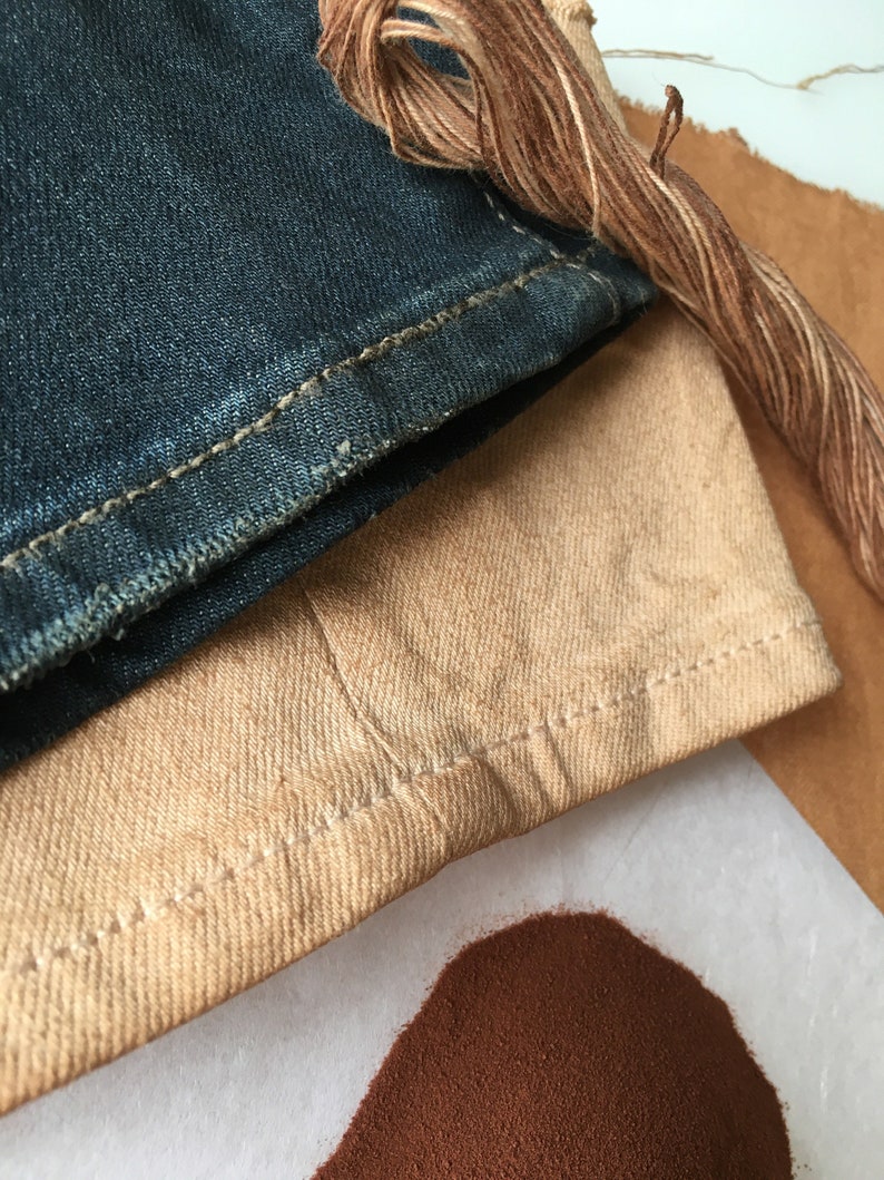 May include: A close-up of a pile of brown dye powder next to a piece of tan fabric and a piece of blue denim fabric. A brown thread is also visible in the image.