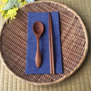 May include: A wooden spoon and chopsticks are placed on a blue and white patterned napkin. The napkin is on a round, woven, brown tray.