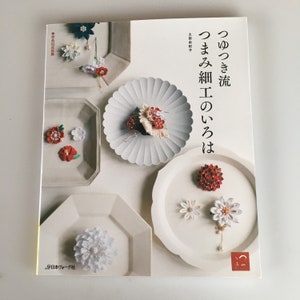 May include: A book titled "Tsuyutsuki-ryu Tsumami Zaiku no Iroha" featuring Japanese fabric flower crafts. The cover displays various fabric flower designs in red, white, and blue, arranged on white plates. The book is published by Nihon Vogue.
