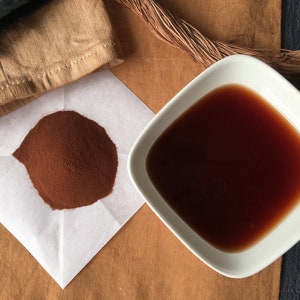 May include: A white square bowl filled with a dark brown liquid, a small pile of brown powder on a white paper, and a brown fabric in the background.