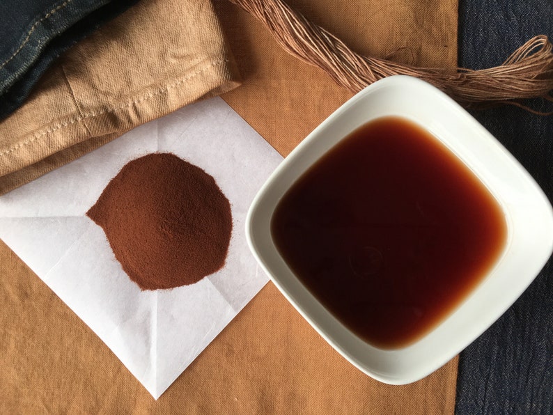 May include: A white square bowl filled with a dark brown liquid, a small pile of brown powder on a white paper, and a brown fabric in the background.