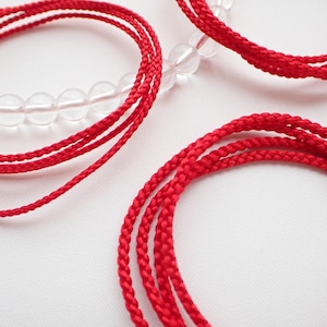 May include: Close-up of red braided cords and clear crystal bead bracelets against a white background. The cords are tightly woven, and the beads are round and translucent. The image highlights the texture and color of the materials.