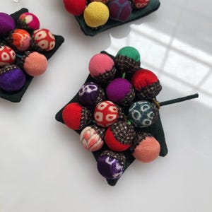 Op de afbeelding: Een cluster van kleurrijke stoffen ballen, elk met een ander patroon, zijn gerangschikt op een zwart vierkant kussen. De ballen zijn met elkaar verbonden door een dun zwart draad.