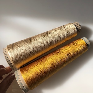May include: Two spools of metallic thread in gold and yellow-gold. The spools are cylindrical with a cardboard base and are filled with shiny, tightly wound thread. The thread has a subtle sheen, reflecting light. The spools are positioned diagonally.
