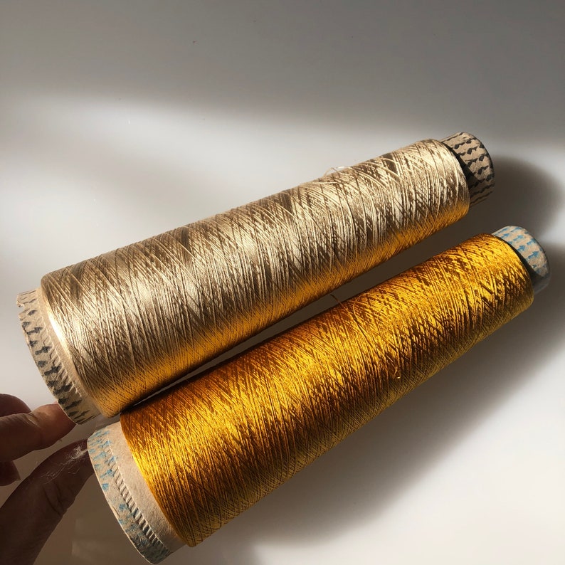 May include: Two spools of metallic thread in gold and yellow-gold. The spools are cylindrical with a cardboard base and are filled with shiny, tightly wound thread. The thread has a subtle sheen, reflecting light. The spools are positioned diagonally.