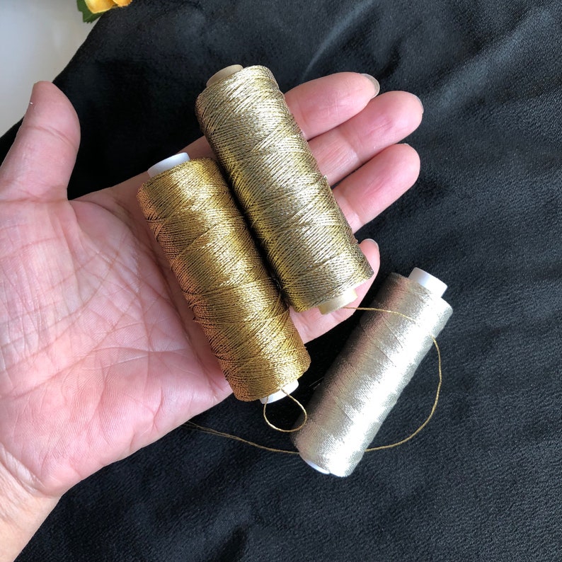 Pu&ograve; includere: Tre bobine di filo metallico, una dorata e due argentate, sono tenute in una mano. Il filo &egrave; avvolto attorno alle bobine ed &egrave; pronto per cucire o ricamare.