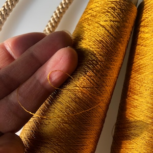 May include: Close-up of golden-yellow spools of thread, with a strand of thread pulled away. The thread is a vibrant, metallic color, and the spools are tightly wound. A gold-colored rope is visible in the background.