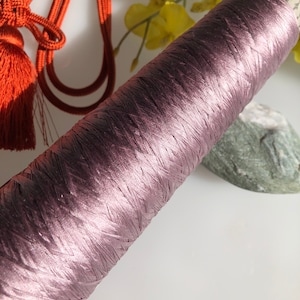 Può includere: Una bobina di filo color malva con una texture lucida e setosa. Il filo è avvolto strettamente, creando una forma cilindrica. Sullo sfondo, un nappa arancione e una pietra verde.