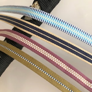 Könnte beinhalten: Vier verschiedene Arten von gewebtem Band mit verschiedenen Farben und Mustern. Die Bänder sind in einem Stapel angeordnet, wobei das oberste Band ein hellblaues und weißes Muster hat, gefolgt von einem beige und dunkelblauen Band, dann einem burgunderfarbenen und weißen Band und schließlich einem dunkelgrünen und weißen Band.