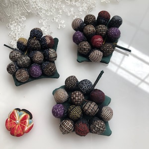 Peut inclure: Trois coussinets à épingles recouverts de tissu vert avec un groupe de boules de tissu colorées sur le dessus. Les boules de tissu sont dans différentes nuances de brun, violet, rouge et noir. Une petite fleur en tissu rouge et jaune avec un centre blanc est également représentée.