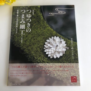May include: A book cover with a white flower made of fabric on a green moss background. The book title is "つまみ細工" in Japanese, which translates to "Tsumami Zaiku" or "Fabric Flower Craft". The book subtitle is "つゆつきの" in Japanese, which translates to "With Dewdrops". The book is about learning how to make fabric flowers.