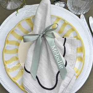 Peut inclure: Une mise en place élégante avec une assiette blanche à rayures jaunes et bord festonné. Une serviette en lin blanche pliée est nouée avec un ruban gris, avec une étiquette nominative "Mélodie". La coutellerie et la verrerie complètent le tout.