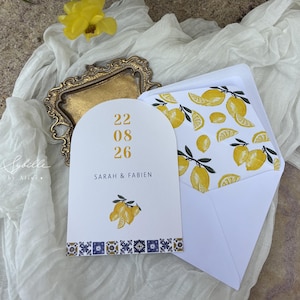 May include: Wedding invitation suite featuring a white arched card with the date "22 08 26" and names "SARAH & FABIEN" in gold text. The card is decorated with lemon illustrations and a blue and white tile border. The envelope has a lemon pattern.