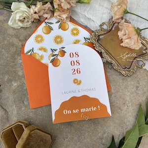 May include: An orange wedding invitation with the text "On se marie!" and the date 08/08/26. The invitation features orange slice and orange branch illustrations. An orange envelope with orange slice illustrations is visible.