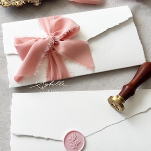 May include: Two white envelopes with deckled edges, one tied with a dusty pink frayed silk ribbon and the other sealed with a round pink wax seal featuring a botanical sprig design. A wooden wax seal stamp and a ring box are also present.