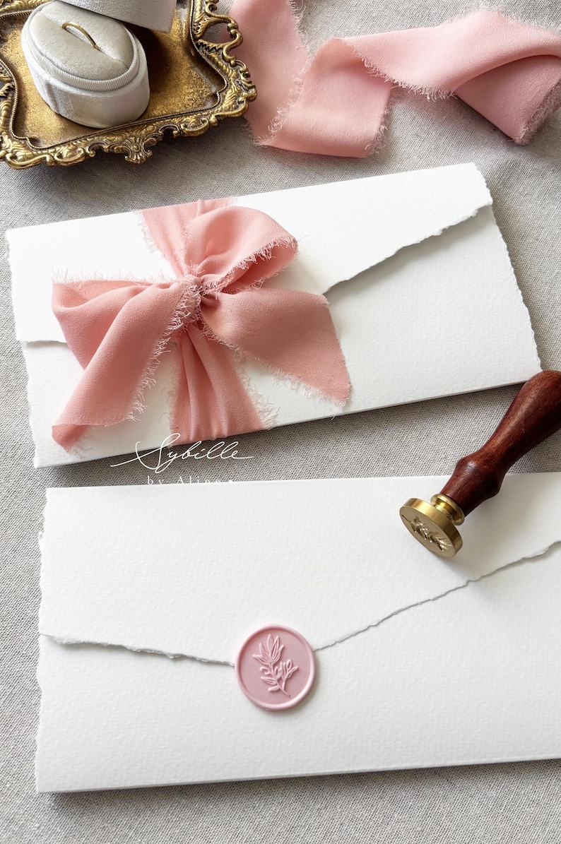 May include: Two white envelopes with deckled edges, one tied with a dusty pink frayed silk ribbon and the other sealed with a round pink wax seal featuring a botanical sprig design. A wooden wax seal stamp and a ring box are also present.