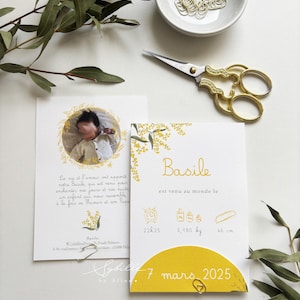 May include: Two baby announcement cards with a photo and text in French, featuring a yellow and white color scheme. The cards include the baby's name, birth details, and the date March 7, 2025. Gold scissors and paperclips are also visible.