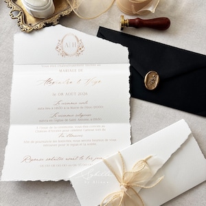 May include: A wedding invitation suite featuring a deckled-edge paper card with gold foil text, a black envelope with a gold wax seal, a wooden wax stamp, a spool of sheer beige ribbon, and a small white ring box on an ornate gold tray.
