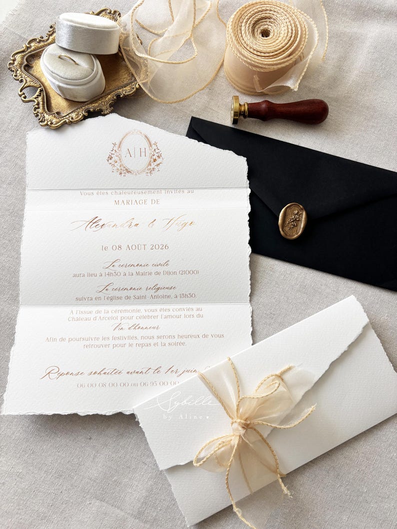 May include: A wedding invitation suite featuring a deckled-edge paper card with gold foil text, a black envelope with a gold wax seal, a wooden wax stamp, a spool of sheer beige ribbon, and a small white ring box on an ornate gold tray.
