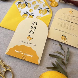 May include: A wedding invitation suite featuring a yellow envelope with a lemon-patterned interior, and matching cards. The cards include the names "LUCIA & EMILIO", the date "23 09 25", and the phrase "that's amore". Gold wax seals and a lemon are also present.