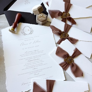 May include: A collection of white wedding invitations with deckled edges, tied with brown chiffon ribbons and secured with gold wax seals. Includes a wooden wax seal stamp, a black envelope with white card, and a beige velvet ring box with two gold rings.
