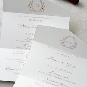 May include: Two white wedding invitation cards with deckled edges, featuring gold and rose gold foil lettering and floral monograms. One card displays the initials A|H, the other L|G. A wooden wax seal stamp and a ring box are visible in the background.