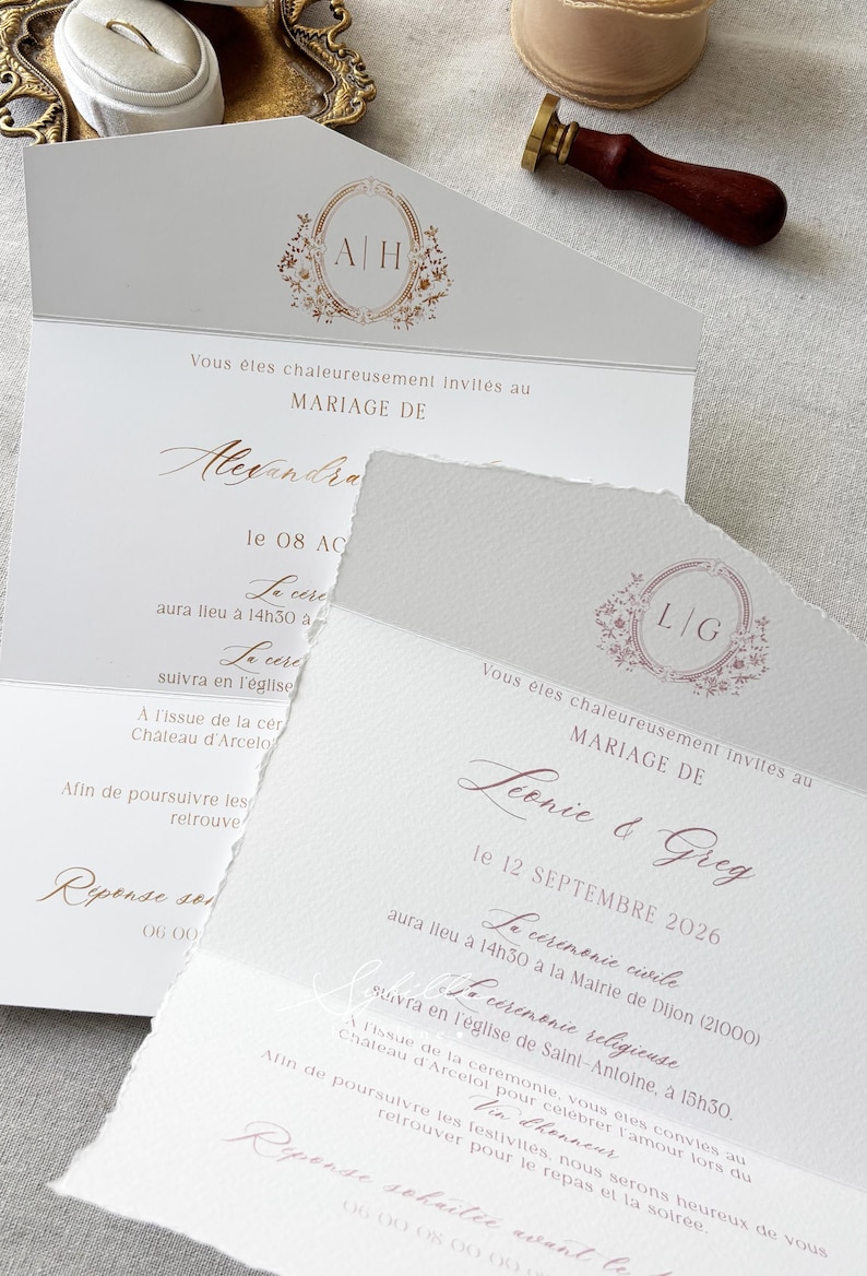May include: Two white wedding invitation cards with deckled edges, featuring gold and rose gold foil lettering and floral monograms. One card displays the initials A|H, the other L|G. A wooden wax seal stamp and a ring box are visible in the background.