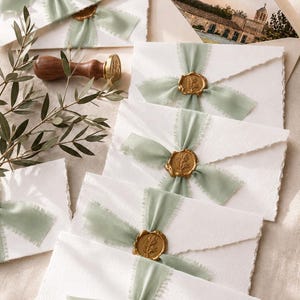 May include: White envelopes with deckled edges, each tied with a sage green frayed silk ribbon and secured with a gold wax seal featuring a botanical design. A wooden wax seal stamp and a photograph of a stone building are also visible.