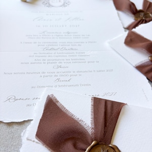 May include: White wedding invitation cards with deckled edges, featuring elegant black script text, brown silk ribbons, and gold wax seals with a botanical leaf design. A wooden wax seal stamp and a small beige ring box are also visible.